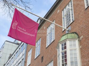 Rosa fasadflagga med texten Svenska Läkaresällskapet monterad på fasadflaggstång vid entrén till Svenska Läkaresällskapets byggnad på Klara Östra Kyrkogata i Stockholm.