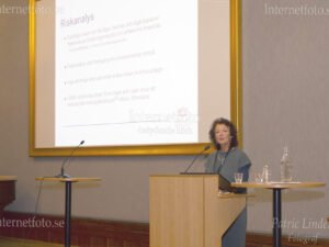 Barnläkare Diana Bornstein-Welff från Mälarsjukhuset i Eskilstuna presenterar en riskanalys vid en läkarkonferens om sjukhushundar och vård. Bilden visar barnläkaren stående bakom en talarstol framför en presentationsduk med information om riskanalys. Mikrofoner glas och vattenkaraff står på bord.