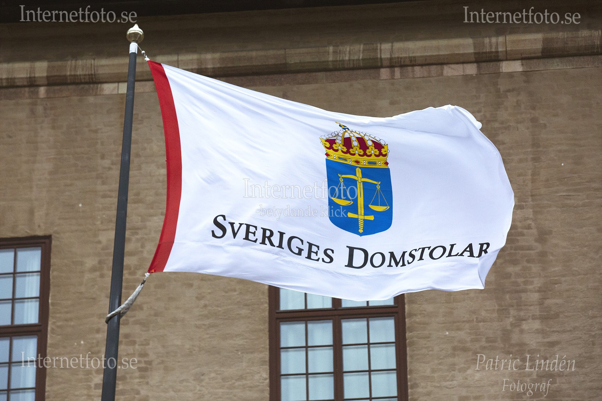 Produktbild endast redaktionell användning. Sveriges Domstolars vita flagga med röd kant, blå sköld och ett krönt emblem gul våg med vågskålar och svärd. Flaggan är sträckt i vinden och vajar från en svart flaggstång framför Stockholms tingsrätt med spröjsade fönster. Texten Sveriges Domstolar syns på flaggan. Bilden är tagen vid Stockholms tingsrätt, Scheelegatan 7, Stockholm, Sverige.