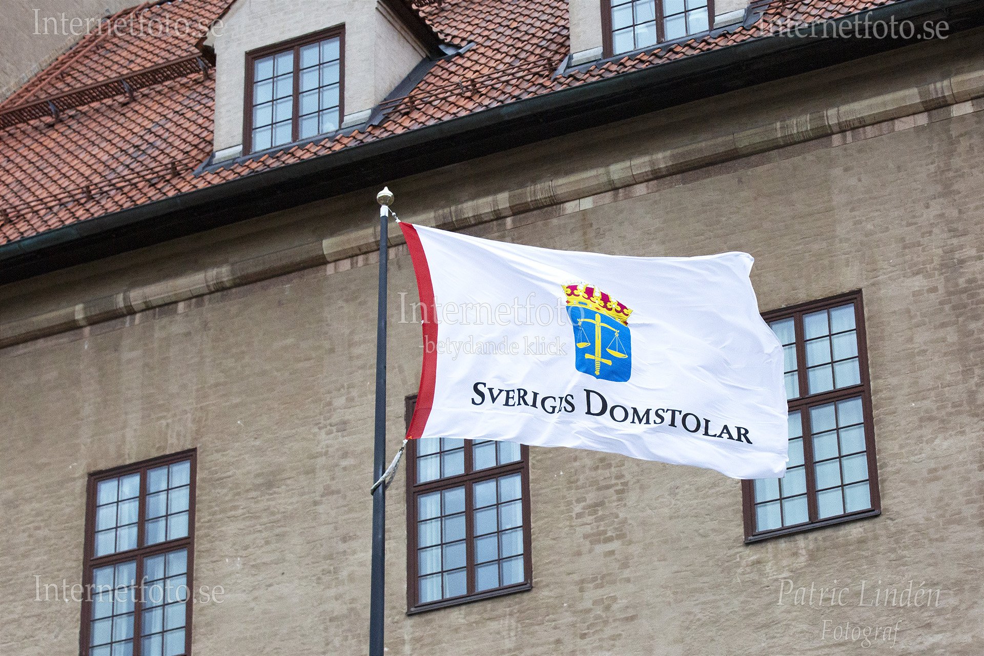 Sveriges Domstolars vita flagga med röd kant, krönt emblem och texten Sveriges Domstolar vajar från en svart flaggstång framför Stockholms tingsrätt på Scheelegatan 7 i Stockholm. Byggnadens tegelfasad har mörkbruna fönster med spröjs samt tak med röda tegelpannor. Bilden är en genrebild med anknytning till svenskt rättsväsende.